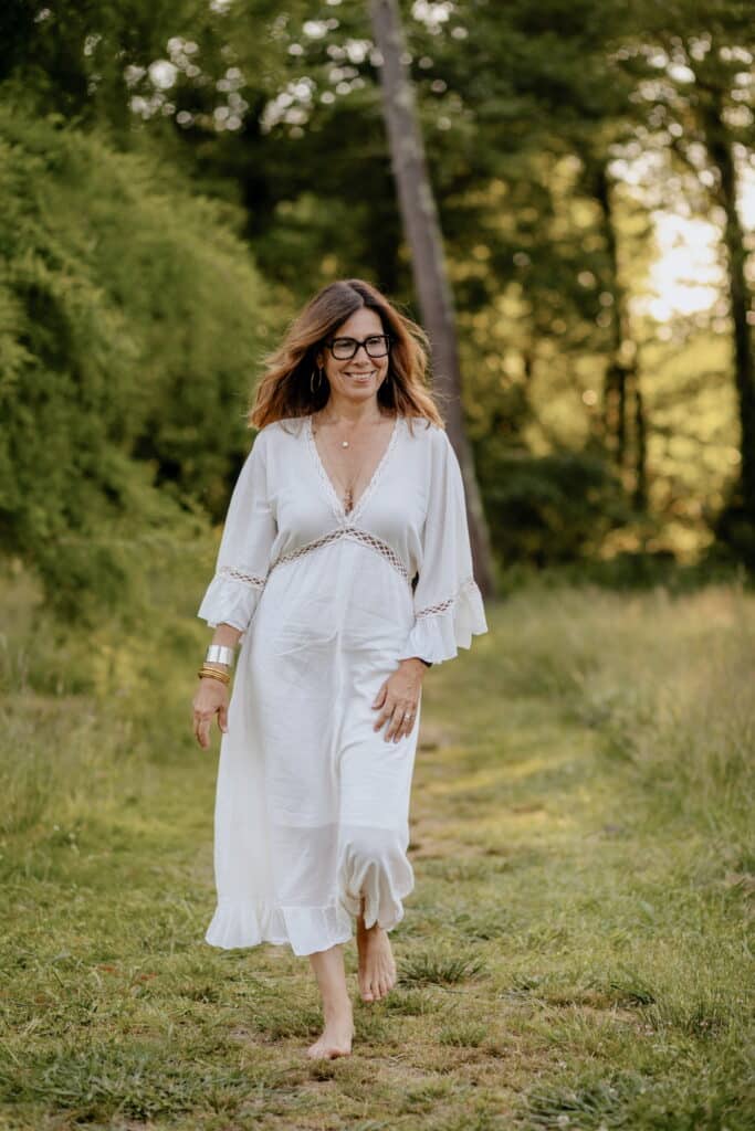 Femme souriante en robe blanche fluide, marchant pieds nus sur un chemin herbeux. Arbres verdoyants, lumière douce.