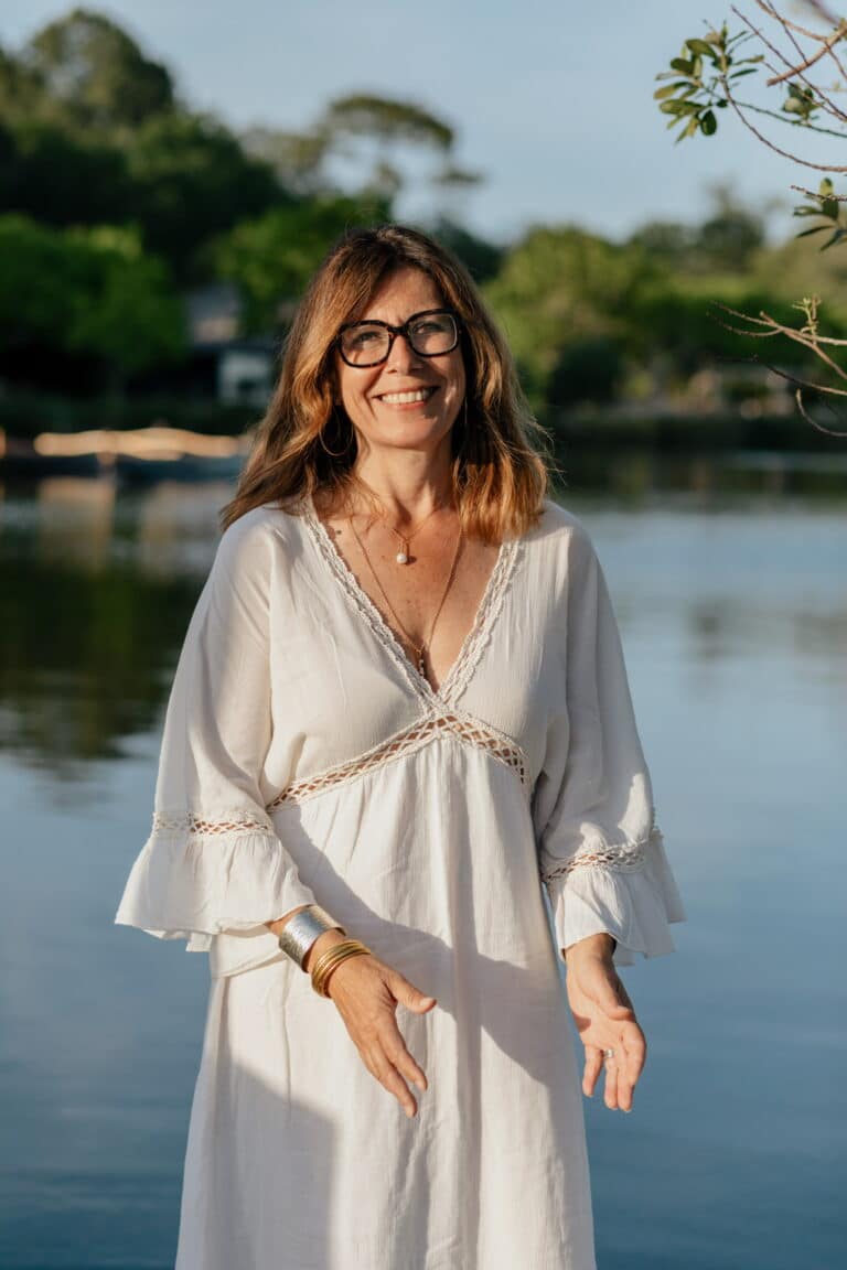 Femme souriante aux lunettes et robe blanche décontractée, posant au bord d'un lac ensoleillé avec nature verdoyante.