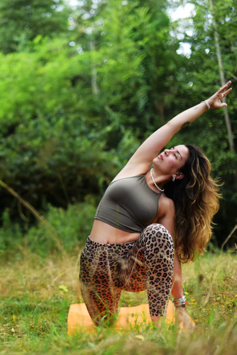 Femme en top vert et legging léopard pratiquant le yoga en nature, un bras levé, sur tapis orange. Vue apaisante.