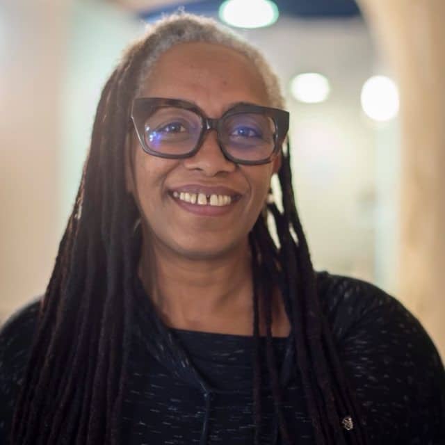 Femme aux dreadlocks grises et lunettes, souriant chaleureusement avec un écart entre les dents de devant, dans un intérieur lumineux.