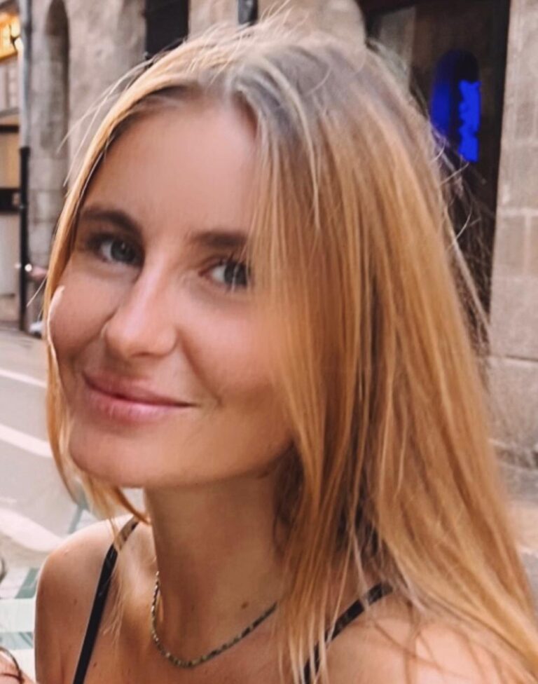 Jeune femme blonde souriante, yeux clairs, portant un collier, en extérieur urbain avec fond flou.
